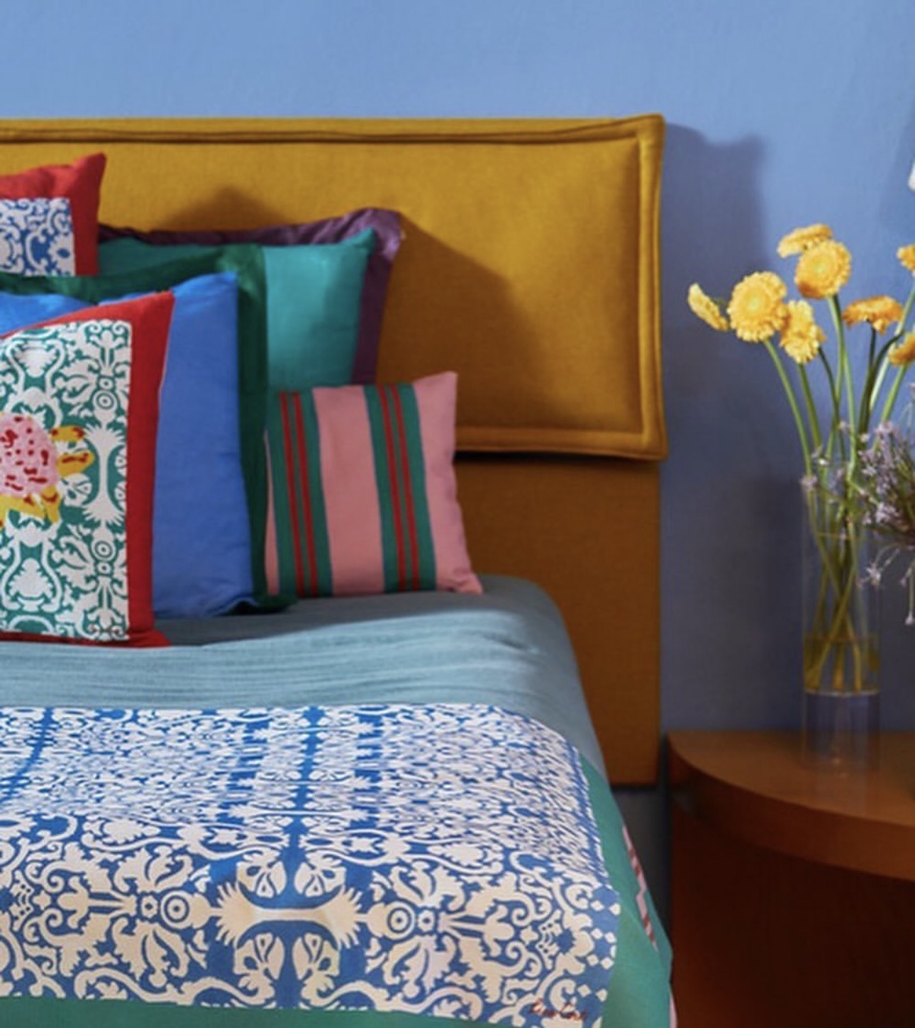 Bed with a mustard headboard, cornflower walls, and patterned Italian linens